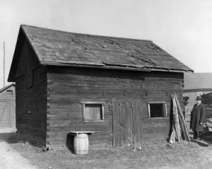 Vincent Roy Jr. storage building, circa 1933. The following is a statement by John A. Bardon of Superior accompanying the photography, "Small storehouse building erected by the late Vincent Roy at Old Superior. The timbers are 4' x 8'. After the one mile dike across Superior Bay had served its purpose, it was allowed to gradually go to pieces. The timbers floating in the Bay for a while were a menace to navigation. You would find them drifting when least expected. The U.S. War Department caused the building of this dike from the end of Rice's Point, straight across to Minnesota Point to prevent the waters of the St. Louis River being diverted from the natural entry at Superior, to the newly dug canal, across Minnesota Point in Duluth. The contention was that, if the waters of the St. Louis were diverted, the natural entrance at Superior would become shoaled from lack of the rivers scouring current. However, when the piers were extended into 18 feet of water at both the old entrance and the Duluth Canal, it was found that the currents of the river had no serious effect. The dike was never popular and was always in the way of the traffic between Superior and Duluth. Several openings were made in it to allow the passage of smaller boats. It was finally condemned by the Government Engineers as a menace to navigation. This all happened in the early 70's. This building is now the only authentic evidence of the dike. It is owned by the Superior and Douglas County Historical Society. The writer is the man in the picture." ~ Wisconsin Historical Society